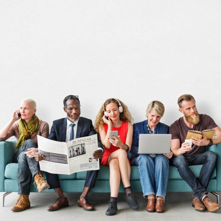 A group of diverse people sit on a couch as they engage with content in a variety of different ways, including by phone, laptop, audio, and newspaper.