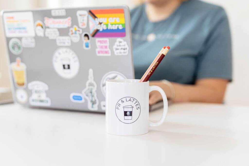 Matisse is in the background working away on her computer. The focus is on a white mug with the PR and Lattes logo that's holding three different coloured pencils with PR and Lattes inscribed on them.