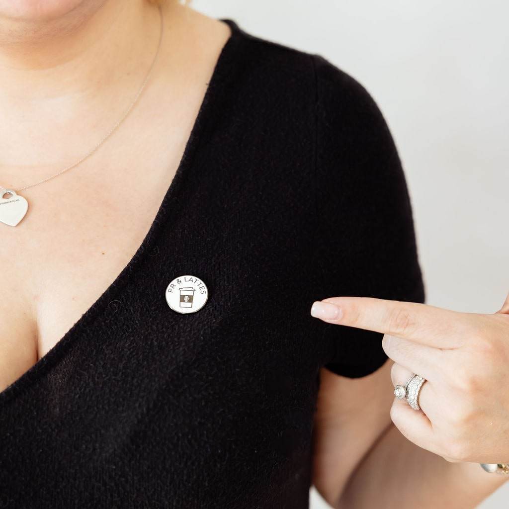 A closeup of the white enamel pin with the PR and Lattes branding. The pin is on Matisse's black top, and she's pointing to it.