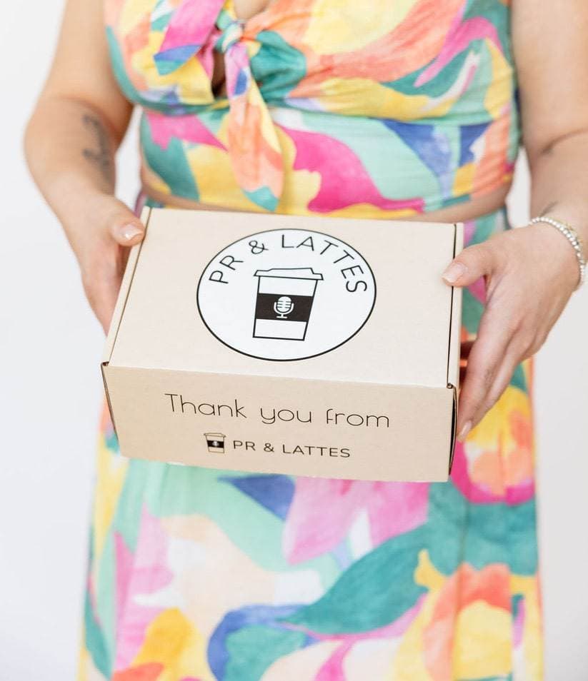 Matisse wears a colourful dress as she holds a light brown box that has the circular PR and Lattes logo on the top, and the text "Thank you from PR & Lattes" on the front fold.