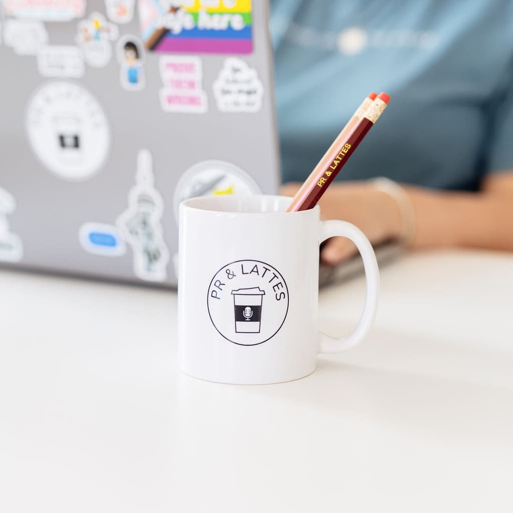 A white mug with the circular PR and Lattes logo holds three differently coloured pencils that are also branded with PR and Lattes. It is sitting in front of a laptop.