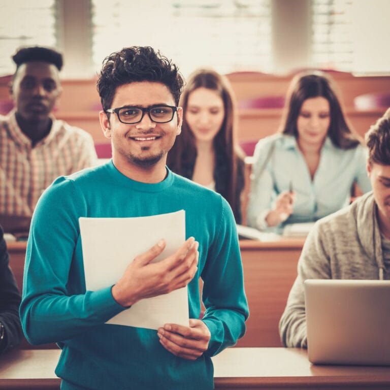 A group of students sit in a lecture hall, with a student of colour standing in the foreground holding a notebook and smiling.