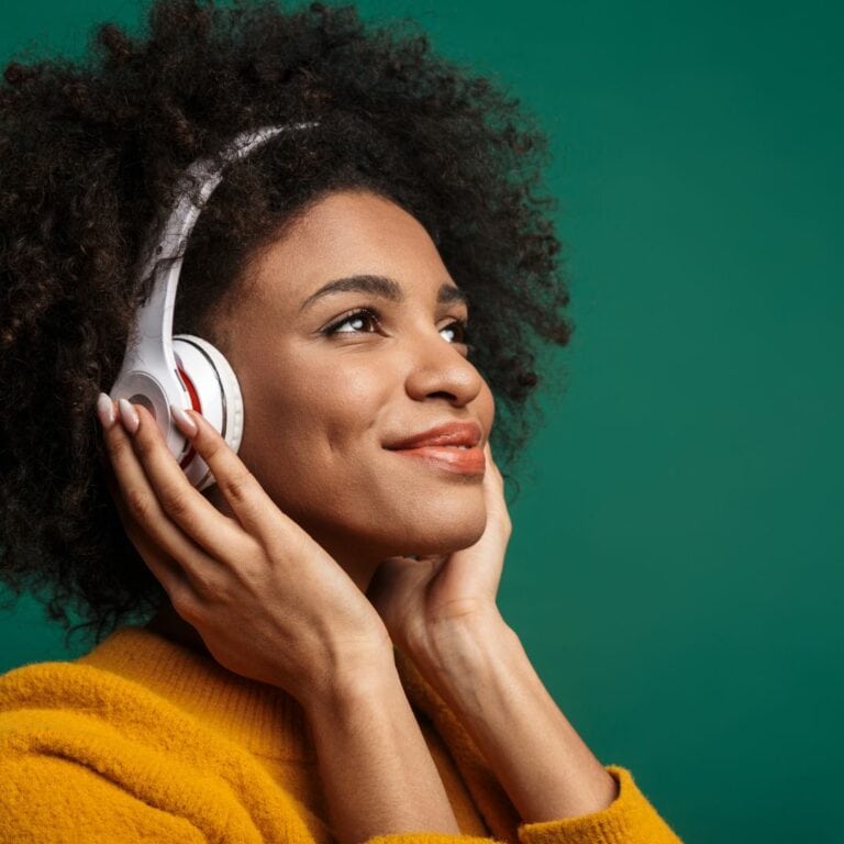 A woman standing in front of a green wall smiles as she listens to music on her white headphones.