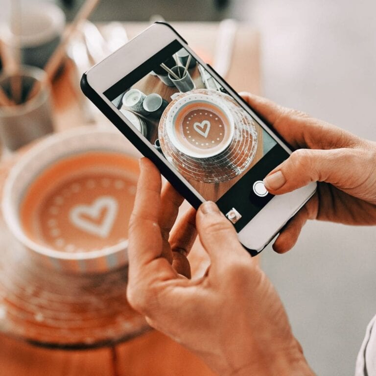 Someone using their smartphone to take a photo of a coffee mug that has coffee and a foam heart in it.