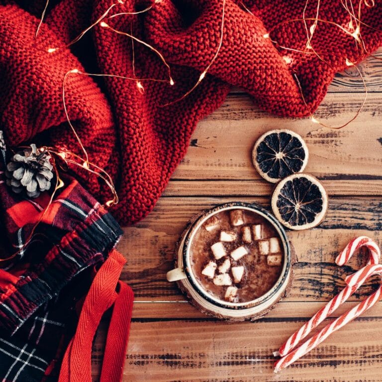 Overhead view of a table with a red blanket, some twinkle lights, a mug filled with hot chocolate and floating marshmallows, and some candy canes.