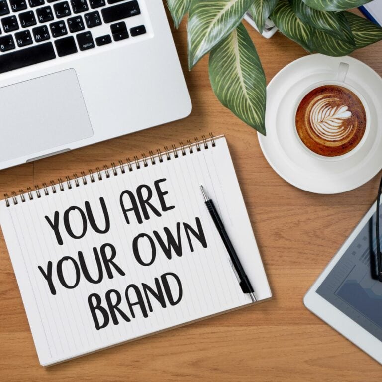 A flat lay display of a laptop, a filled coffee cup, a tablet, and a notepad with the text, "You are your own brand."