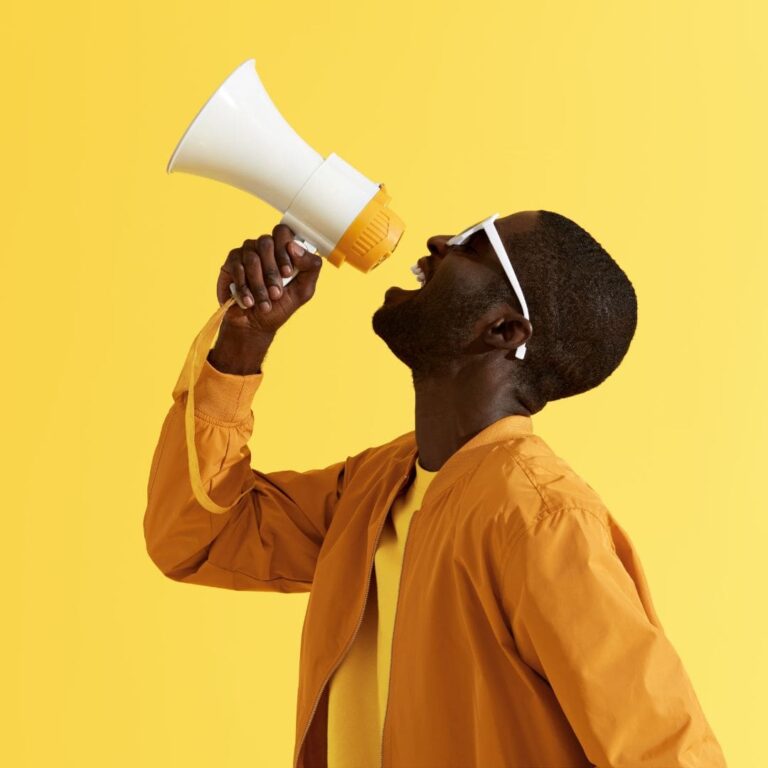 A young Black man wearing all yellow is shouting into a megaphone on a yellow background.