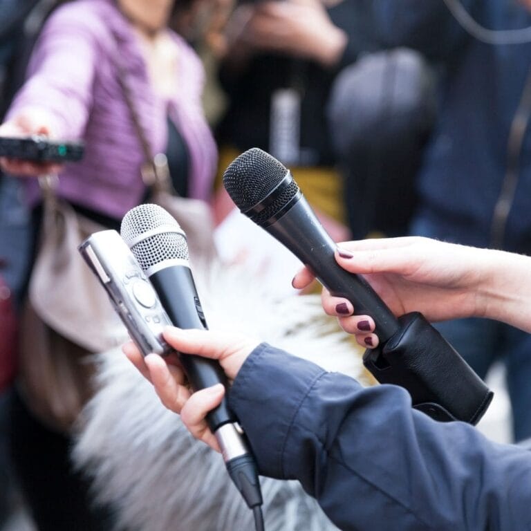 Two hands are holding microphones and recorders in front of a crowd.
