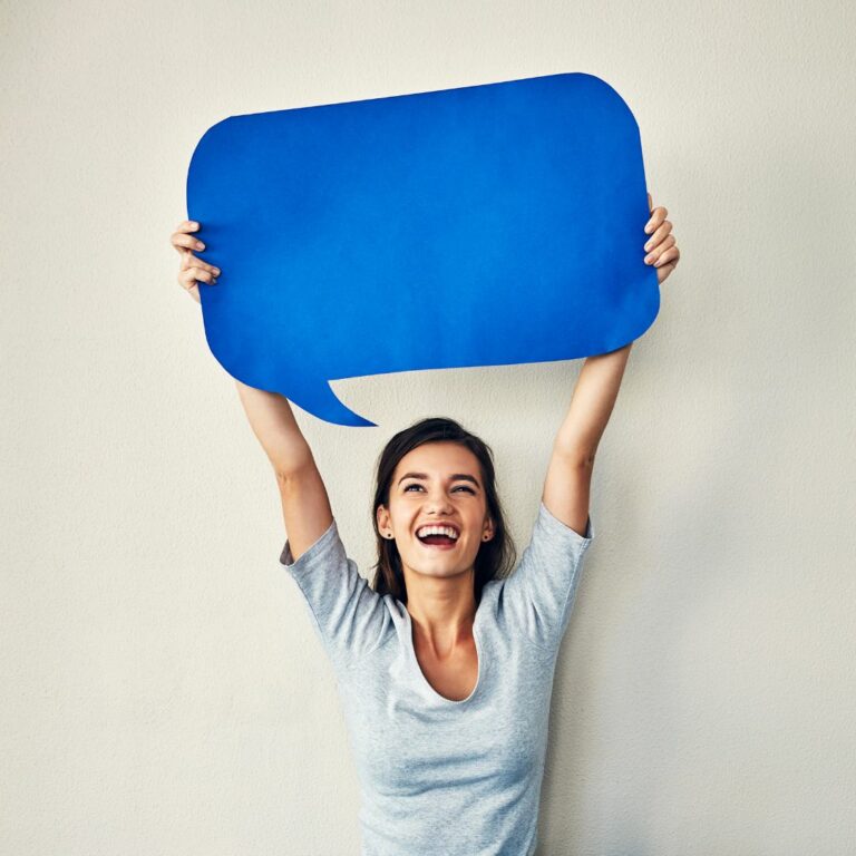 A woman in a grey shirt holds up a large blue conversation bubble made of cardboard above her head.